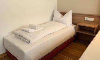 A simple bedroom with a single bed, fresh towels, and a bedside table. The walls are light and the floor is made of wood.