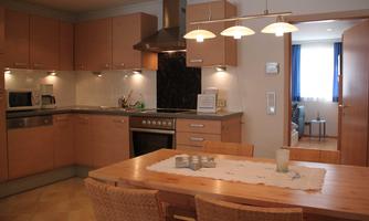 A modern kitchen with wooden cabinets and stainless steel appliances. A wooden table stands in the center, decorated with a tablecloth and some accessories.