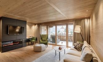A modern living room with a cozy sofa and two green armchairs. Large windows offer a view of the surrounding landscape.