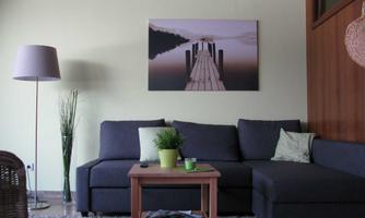 A cozy living room with a gray sofa and a small table. On the wall hangs a picture of a pier over water, and a lamp and plants complete the decor.