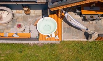 A cozy outdoor area with a wooden terrace, a hot tub, and a dining area. The surroundings are adorned with green grass and elegant stones.