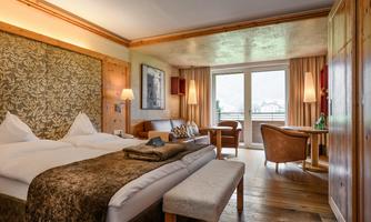 A cozy hotel room with two single beds, a wooden chair, and a small table. Large windows provide plenty of daylight and offer a view outside.