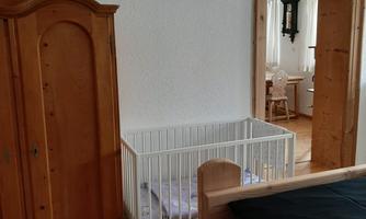 A bright room with a white children's bed and a wooden dresser. In the background, another room with furniture is visible.