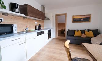 A modern kitchen with white cabinets and wood details. In the living room, there is a gray sofa with cushions and a dining table.