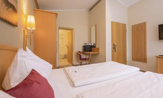 A cozy bedroom with a large bed and a bright, inviting space. In the background, a desk and access to the bathroom are visible.