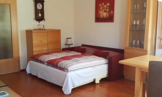 A cozy bedroom with a double bed and a wooden furniture combination. The walls are painted light, and there is a clock as well as a dining table in the room.