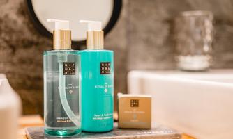 Two bottles of shampoo and shower gel are sitting next to a bar of soap on a bathroom shelf. In the background, a round mirror is visible.