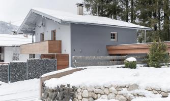 A modern house in the snow with a gray facade paint. Surrounded by snowy trees and a stone wall.