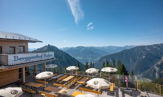 A mountain guesthouse with an outdoor terrace and a view of the surrounding mountains. The sky is blue with some clouds.