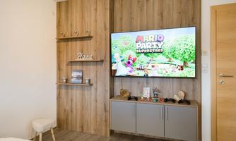 A modern room with a television displaying the game "Mario Party Superstars." In the background, there are wooden shelves and a cozy seating area.