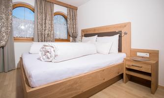 A cozy bedroom with a large wooden bed and soft white bedding. The windows are adorned with elegant curtains, and the room is bright and inviting.