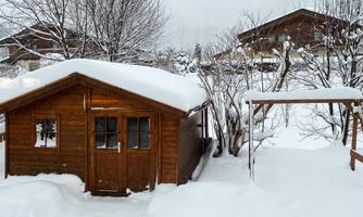 A cozy wooden house, surrounded by deep snow. In the background, there are more buildings and snow-covered trees.
