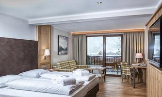 A modern hotel room with a large bed and a cozy sofa. There is a balcony with a view of the landscape and a bright, inviting atmosphere.