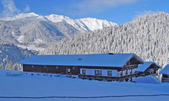 A snowy winter landscape with tall mountains in the background. In the foreground, wooden buildings are surrounded by a thick layer of snow.