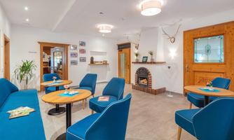 A cozy lounge area with blue chairs and wooden details. A fireplace and plants create an inviting atmosphere.