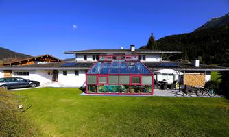 A modern house with a spacious garden and a greenhouse. In the background, mountains and a clear blue sky are visible.
