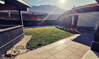 A sunny garden with green grass and a view of the mountains. The terrace is designed with tiles and stones.