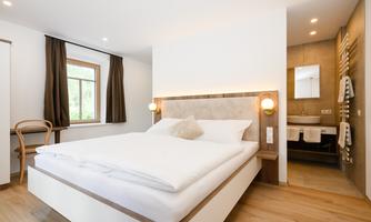 A modern bedroom with a large double bed and light wooden flooring. The windows let in plenty of light, and an adjacent bathroom is visible.