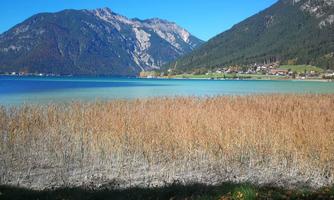 A clear lake surrounded by mountains and golden reeds. The sky is blue, and the day seems to be sunny.