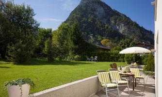 A beautiful garden with comfortable deck chairs and a parasol. In the background, a majestic mountain rises under a clear blue sky.
