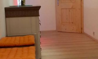 A bright room with a wooden floor and an open door. On the left side, there are two orange cushions on a piece of furniture.