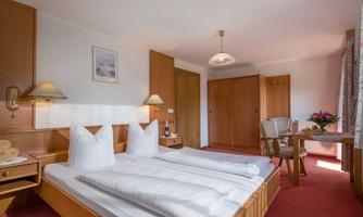 A cozy bedroom with two single beds and wood-colored furnishings. The room features a table, chairs, and a window with curtains.