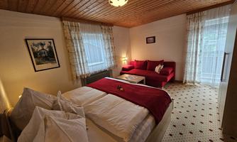 A cozy bedroom with a double bed and red bedding. The room features a red sofa and large windows that let in plenty of light.