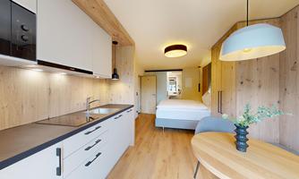 A modern living kitchen with a dining table and a cozy bedroom in the background. The walls are made of light wood, which creates a warm atmosphere.
