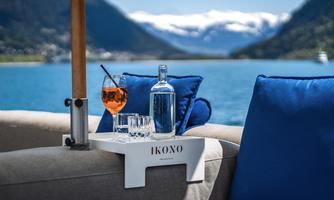 A beverage tray with a glass of cocktail, a bottle of water, and glasses on a couch with blue pillows. In the background, a beautiful lake extends with mountains and a clear sky.