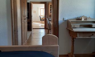A cozy room with a wooden frame bed and a desk. In the background, a passage to another room is visible.