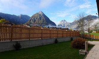 A tranquil landscape with green meadow and a wooden fence. In the background, majestic mountains and a blue sky can be seen.