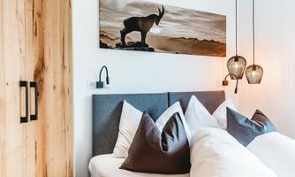 A cozy bedroom with a gray headboard and white pillows. A picture of a mountain animal hangs on the wall, while stylish pendant lights create a warm atmosphere.