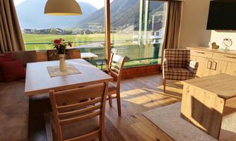 A cozy living room with wooden furniture and a dining area. Large windows provide a view of the mountains and the green landscape.