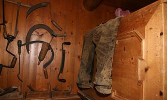A wooden room with agricultural tools on the wall. On a shelf lie an old pair of trousers and a checkered shirt.