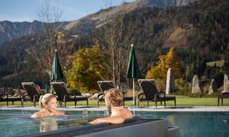 A relaxing outdoor pool with two people, surrounded by mountains and colorful foliage. Umbrellas and sun loungers complete the idyllic scene.