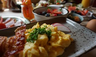 A hearty breakfast with scrambled eggs, bacon, and fresh chives. In the background, fruit variations and other breakfast treats can be seen.