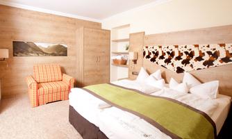 A cozy hotel room with a large bed and colorful pillows. Next to the bed is a checkered armchair, and on the wall hangs a picture of mountains.