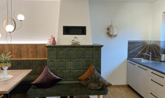 A modern living and dining room with a cozy bench and a red tiled heater. The open kitchen is bright and minimalist.