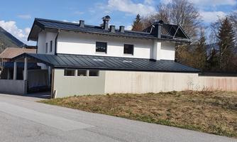 A modern house with a flat roof and a light facade. In the background, trees and mountains are visible.