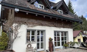 A cozy house with a combination of wooden and white facade. The entrance is surrounded by plants and there are large windows that let in light.