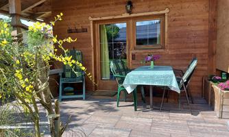 A cozy terrace with a table and chairs in front of a wooden paneling. Flowers in boxes and fresh greenery create an inviting atmosphere.