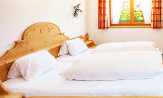 A bright bedroom with a wooden bed and large white pillows. There is a window with red curtains that lets in daylight.