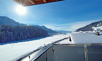 A winter landscape with snow-covered mountains and forests. The sun shines brightly in the blue sky.