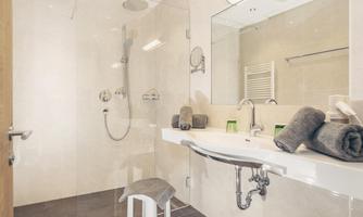 A modern bathroom with a shower and a large mirror. On the sink are gray towels and a stool is ready.