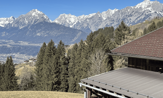 A picturesque mountain landscape with snow-capped peaks and green grass. In the foreground, there is a wooden house with a slanted roof.