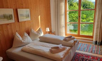 A cozy bedroom with two beds and bright wooden walls. The large window offers a view of a green garden.