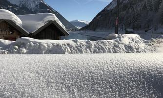 A snowy winter landscape with cabins and high mountains in the background. The sky is clear and blue with some clouds.