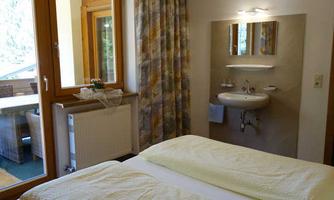 A cozy bedroom with two beds and a sink. Large windows provide plenty of light and a view of a balcony.