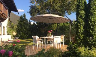 An inviting garden with a dining table and chairs under a sun umbrella. Pink flowers and green trees surround the area.