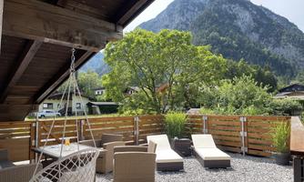 A cozy terrace with loungers and a hanging chair. In the background, green trees and majestic mountains can be seen.